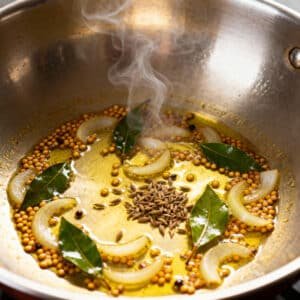 Tempering mustard seeds, curry leaves, and onions in hot ghee inside a steel pan.